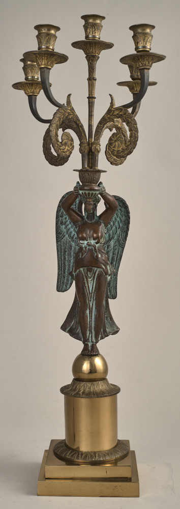 Kandelabr z uskrzydloną postacią na postumencie przymającą w rękach ramiona świecznika.