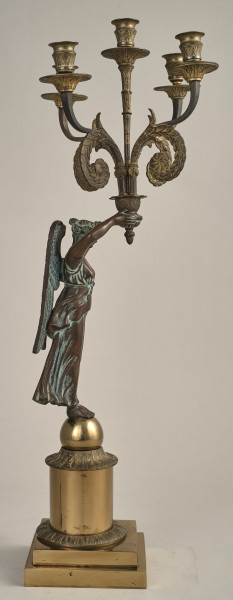 Kandelabr z uskrzydloną postacią na postumencie przymającą w rękach ramiona świecznika.