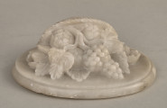 Alabastrowy przycisk do papieru ma kształt owalnego medalionu. Ozdobiony jest półplastyczną kompozycją z kwiatów, gałązek winorośli i owoców umieszczonych w koszu