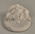 Alabastrowy przycisk do papieru ma kształt owalnego medalionu. Ozdobiony jest półplastyczną kompozycją z kwiatów, gałązek winorośli i owoców umieszczonych w koszu