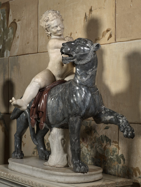 Marmurowa grupa rzeźbiarska przedstawia małego chłopca - Dionizosa siedzącego na grzbiecie pantery. Elementy grupy w różnych kolorach na tle ściany kamiennej. Widok 3/4