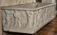 Sarkofag marmurowy z przedstawieniem orszaku dionizyjskiego. Ustawiony na podłodze z dwukolorowego parkietu. Widok od strony wąskiego boku.