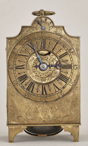 Zegar kominkowy w kształcie prostopadłościanu z centralnie umieszczoną tarczą zegarową z uchwytem do przenoszenia. Zegar złocony z dekoracją reliefową.