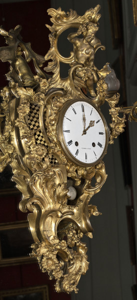 Zegar Cartel. Złocony zegar wiszący na lustrze, bardzo bogato dekorowany motywami roślinnymi. W centralnej części duża biała tarcza zegarowa. Bok lewy