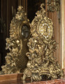 Zegar kominkowy ze złoconego brązu stojący na tle lustra. Zegar  bardzo bogato zdobiony sceną mitologiczną przedstawiającą Narodziny Wenus. Nad sceną złota tarcza zegarowa. Zdjęcie wykonane w ujęciu 3/4 ukazuje odbicie w lustrze tylnej, płaskiej ścianki oraz otwór z widocznym mechanizmem