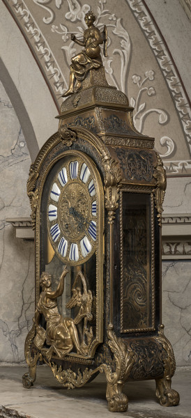 Ujęcie boczne stojącego zegara z mosiężnymi inkrustacjami i złoceniami. W części centralnej złota tarcza z cyframi rzymskimi. W części dolnej i w szczycie przedstawienia figuralne. Widoczna ścianka boczna.
