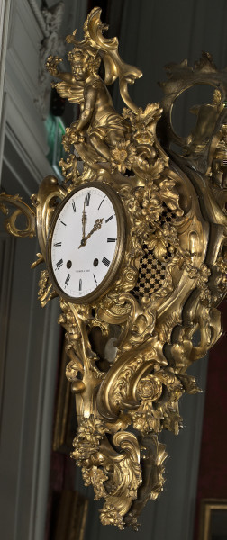 Zegar Cartel. Złocony zegar wiszący na lustrze, bardzo bogato dekorowany motywami roślinnymi. W centralnej części duża biała tarcza zegarowa. Bok prawy