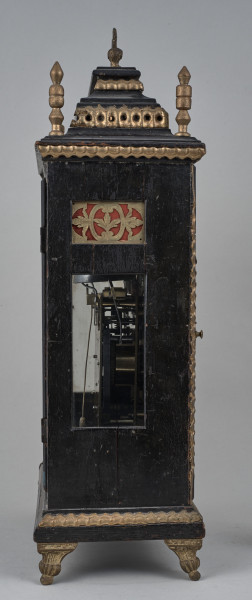 Zegar szafkowy z uchwytem górnym do przenoszenia. Prostopadłościenna obudowa drewniana, czerniona, złocona, posiada aplikacje mosiężne. Ścianka boczna z otworem, przez który widać mechanizm.