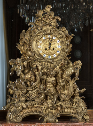Zegar kominkowy ze złoconego brązu stojący na tle lustra. Zegar  bardzo bogato zdobiony sceną mitologiczną przedstawiającą Narodziny Wenus. Nad sceną złota tarcza zegarowa z białymi, okrągłymi miejscami na rzymskie cyfry.