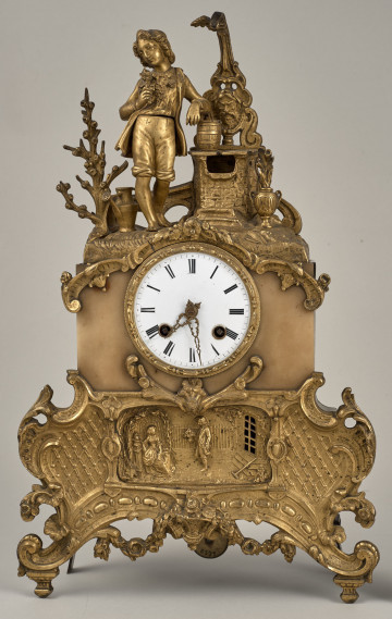 ﻿Zegar kominkowy wykonany z brązu, wsparty na czterech nóżkach, bogato dekorowany z białą tarczą w centralnej części.