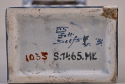 Fragment ceramicznej podstawy zegara z widocznymi znakami producenta oraz numerem inwentarzowym muzeum.