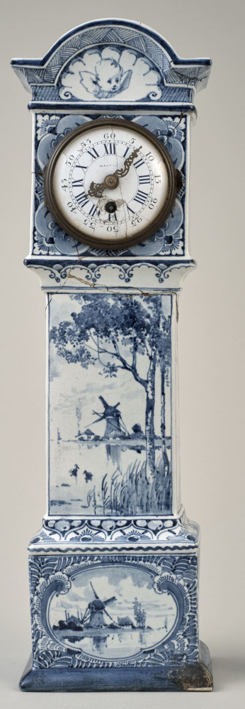 Zegar na białym cokole ceramicznym z niebieskimi holenderskimi przedstawieniami wiatraków w typie Delft. Duża tarcza zegarowa u góry cokołu zwieńczona półokrągłym daszkiem. Ujęcie frontalne
