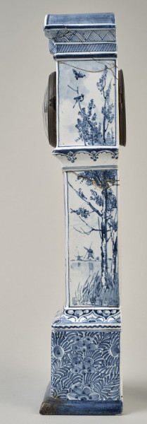 Zegar na białym cokole ceramicznym z niebieskimi holenderskimi przedstawieniami wiatraków w typie Delft.  Bok lewy