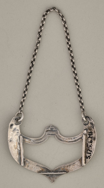 Etykietka srebrna na łańcuszku w kształcie stylizowanego kartusza zakończonego w górnych narożach głową barana (po obu stronach). Odwrocie