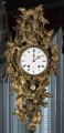 Zegar Cartel. Złocony zegar wiszący na lustrze, bardzo bogato dekorowany motywami roślinnymi. W centralnej części duża biała tarcza zegarowa. Ujęcie frontalne