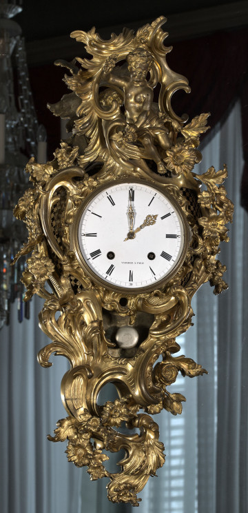 Zegar Cartel. Złocony zegar wiszący na lustrze, bardzo bogato dekorowany motywami roślinnymi. W centralnej części duża biała tarcza zegarowa. Ujęcie frontalne