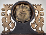 Fragment tylnej strony zegara szafkowego z widocznym mechanizmem i dekoracją roślinną w kolorze złotym