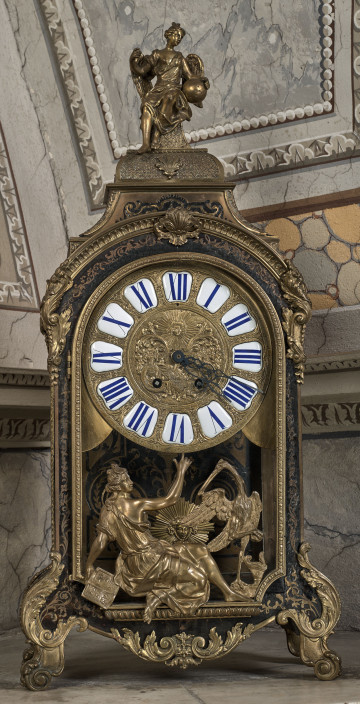 Przód stojącego zegara z mosiężnymi inkrustacjami i złoceniami. W części centralnej złota tarcza z cyframi rzymskimi. W części dolnej i w szczycie przedstawienia figuralne.