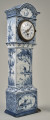 Zegar na białym cokole ceramicznym z niebieskimi holenderskimi przedstawieniami wiatraków w typie Delft. Duża tarcza zegarowa u góry cokołu zwieńczona półokrągłym daszkiem. Ujęcie 3/4