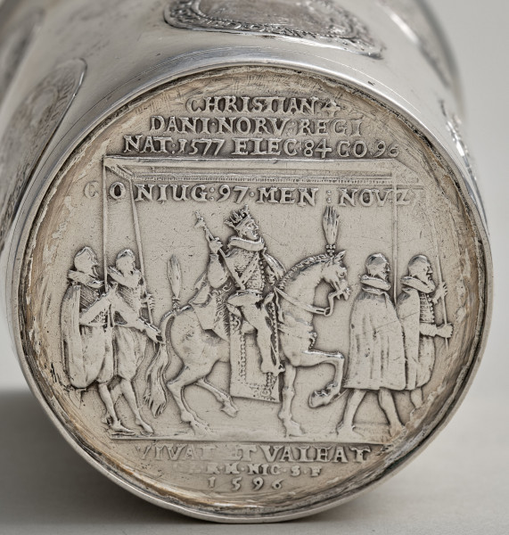 Spodnia część dna srebrnego kubka z przedstawieniem króla na koniu i czterech osób niosących baldachim. Widoczne łacińskie napisy z datą 1596 u dołu.