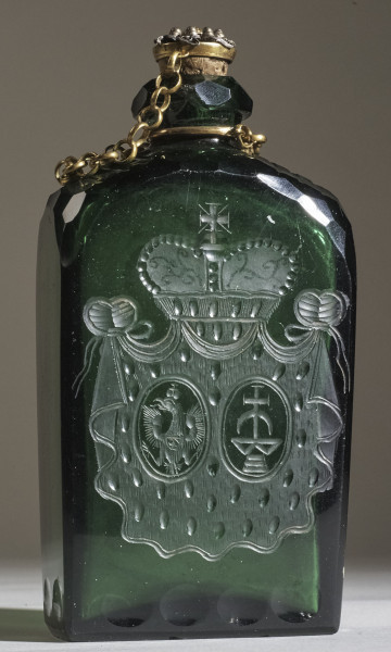 Flasza z zielonego szkła z herbem Trąby i Waga. Widoczny złoty łańcuszek z korkiem.