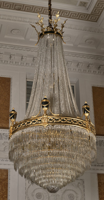 Żyrandol kryształowy w kształcie gruszki ze złotą obręczą dekorowaną urnami na tle ściany i sufitu