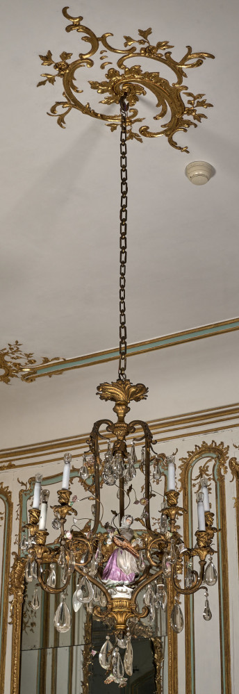 Żyrandol wieloświecowy, wiszący na długim łańcuchu ozdobiony figurką porcelanową dziewczyny. Na tle sufitu i bogato dekorowanej ściany z lustrem