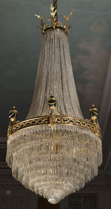 Żyrandol kryształowy w kształcie gruszki ze złotą obręczą dekorowaną urnami na tle malowanego w chmury sufitu