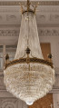 Żyrandol kryształowy w kształcie gruszki ze złotą obręczą dekorowaną urnami na tle ściany