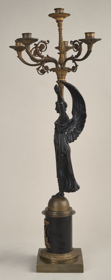 Kandelabr 5 świecowy, składa się z podstawy - cokołu, trzonu w postaci stojącej uskrzydlonej Nike, która wzniesionymi do góry rękami podtrzymuje nastawę z pięcioma wygiętymi ramionami na świece. Bok prawy