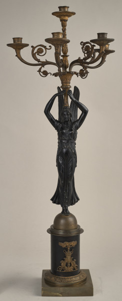 Kandelabr 5 świecowy, składa się z podstawy - cokołu, trzonu w postaci stojącej uskrzydlonej Nike, która wzniesionymi do góry rękami podtrzymuje nastawę z pięcioma wygiętymi ramionami na świece.