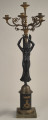 Kandelabr 5 świecowy, składa się z podstawy - cokołu, trzonu w postaci stojącej uskrzydlonej Nike, która wzniesionymi do góry rękami podtrzymuje nastawę z pięcioma wygiętymi ramionami na świece.