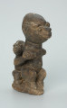 rzeźba; przedmiot obrzędowy; Figura kultu sił wegetacji - Ujęcie z przodu z prawej strony. Rzeźbiona w grafitowym steatycie siedząca postać ludzka o cechach kobiecych z podkurczonymi nogami i dzieckiem na plecach.