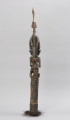 flet obrzędowy - Ujęcie tyłu; Podłużny, bogato zdobiony flet wykonany z odcinka bambusa otwartego po stronie służącej do zadęcia i zamkniętego naturalnym kolankiem po stronie przeciwnej, gdzie znajduje się drewniana figura kobiety-przodka w pozycji klęczącej z ptakiem na głowie.
