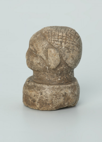 rzeźba; przedmiot obrzędowy; Głowa - Ujęcie lewy bok. Rzeźbiona w szarobeżowym steatycie głowa ludzka na okrągłej podstawie z charakterystycznym uczesaniem lub nakryciem głowy w formie płaskiego ornamentu kostkowego.