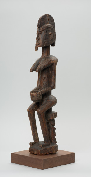 drewniana figura - Ujęcie z przodu, z lewej strony. Drewniana, rzeźbiona figura kobiety, siedzącej na stołku.
