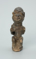 rzeźba; przedmiot obrzędowy; Figura kultu sił wegetacji - Ujęcie z przodu. Rzeźbiona w grafitowym steatycie siedząca postać ludzka o cechach kobiecych z podkurczonymi nogami i dzieckiem na plecach.