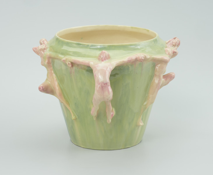 ceramika - Ujęcie z kolejnego boku; Na wprost widoczna przyczepiona na zielonym wazonie postać meska wygieta do tyłu, zaczepiona nogami i głową o wazon, trzymająca po bokach postacie za ręce.
