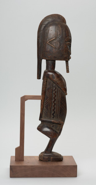 figura - postać przodka - Ujęcie z boku z prawej strony. Drewniana rzeźba kobiety o zgeometryzowanej formie zdobiona licznymi grupami nacięć. Sylwetka spłaszczona po bokach.