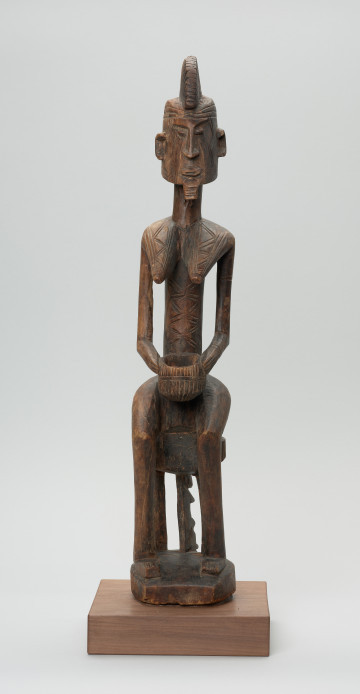 drewniana figura - Ujęcie z przodu. Drewniana, rzeźbiona figura kobiety, siedzącej na stołku.