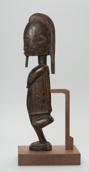 figura - postać przodka - Ujęcie z boku z lewej strony. Drewniana rzeźba kobiety o zgeometryzowanej formie zdobiona licznymi grupami nacięć. Sylwetka spłaszczona po bokach.