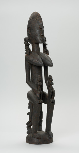 drewniana figura - Ujęcie z przodu, z lewej strony. Drewniana, rzeźbiona figura kobiety. Na jej ramionach i rękach znajdują się mniejsze figurki.