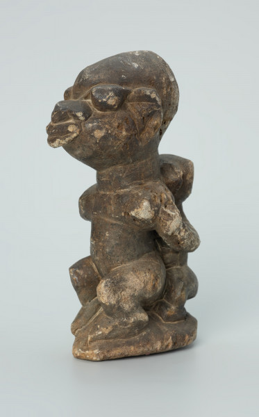 rzeźba; przedmiot obrzędowy; Figura kultu sił wegetacji - Ujęcie z przodu z lewej strony. Rzeźbiona w grafitowym steatycie siedząca postać ludzka o cechach kobiecych z podkurczonymi nogami i dzieckiem na plecach.