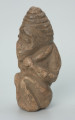 rzeźba; przedmiot obrzędowy; Figura kultu zmarłych - Ujęcie z przodu z lewej strony. Rzeźbiona w szarobeżowym steatycie postać ludzka ze spiralną linią na czubku dużej głowy, siedząca z przykurczonymi nogami i trzymająca w dłoniach bliżej nieokreślone przedmioty, kształtem przypominające motyki.
