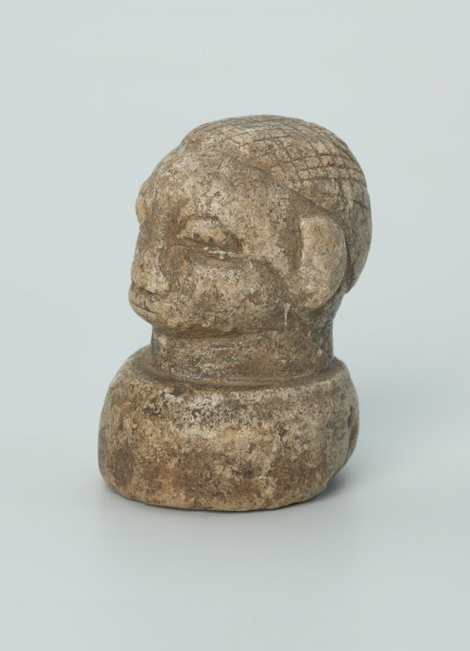 rzeźba; przedmiot obrzędowy; Głowa - Ujęcie z przodu z lewej strony. Rzeźbiona w szarobeżowym steatycie głowa ludzka na okrągłej podstawie z charakterystycznym uczesaniem lub nakryciem głowy w formie płaskiego ornamentu kostkowego.
