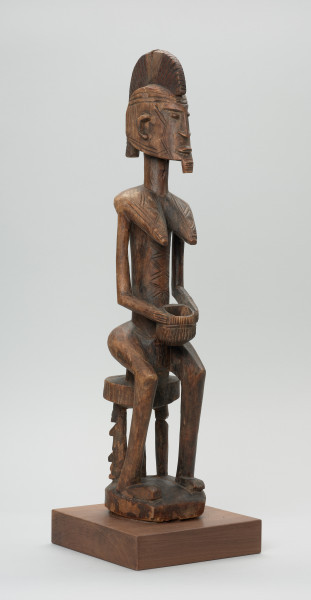 drewniana figura - Ujęcie z przodu, z prawej strony. Drewniana, rzeźbiona figura kobiety, siedzącej na stołku.