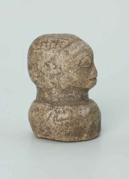 rzeźba; przedmiot obrzędowy; Głowa - Ujęcie prawy bok. Rzeźbiona w szarobeżowym steatycie głowa ludzka na okrągłej podstawie z charakterystycznym uczesaniem lub nakryciem głowy w formie płaskiego ornamentu kostkowego.