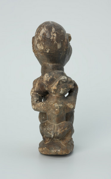 rzeźba; przedmiot obrzędowy; Figura kultu sił wegetacji - Ujęcie z tyłu. Rzeźbiona w grafitowym steatycie siedząca postać ludzka o cechach kobiecych z podkurczonymi nogami i dzieckiem na plecach.