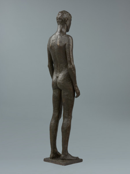 akt męski - Ujęcie z tyłu skosem w prawą stronę; Rzeźba z brązu przedstawiająca nagiego, młodego, szczupłego mężczyznę, stojącego twarzą do widza z rękami wzdłuż ciała.