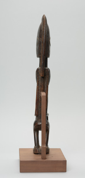 figura - postać przodka - Ujęcie z tyłu. Drewniana rzeźba kobiety o zgeometryzowanej formie zdobiona licznymi grupami nacięć. Sylwetka spłaszczona po bokach.